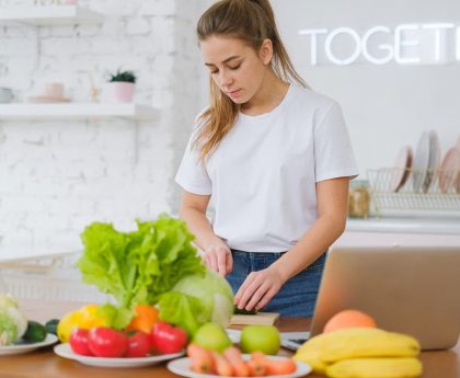 Woman in white t-shirt preparing fresh vegetables in a bright kitchen with a laptop.