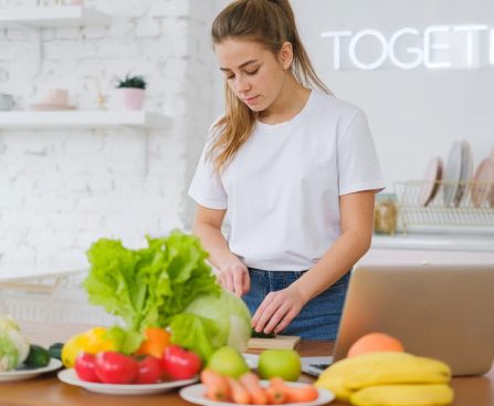 Woman in white t-shirt preparing fresh vegetables in a bright kitchen with a laptop.